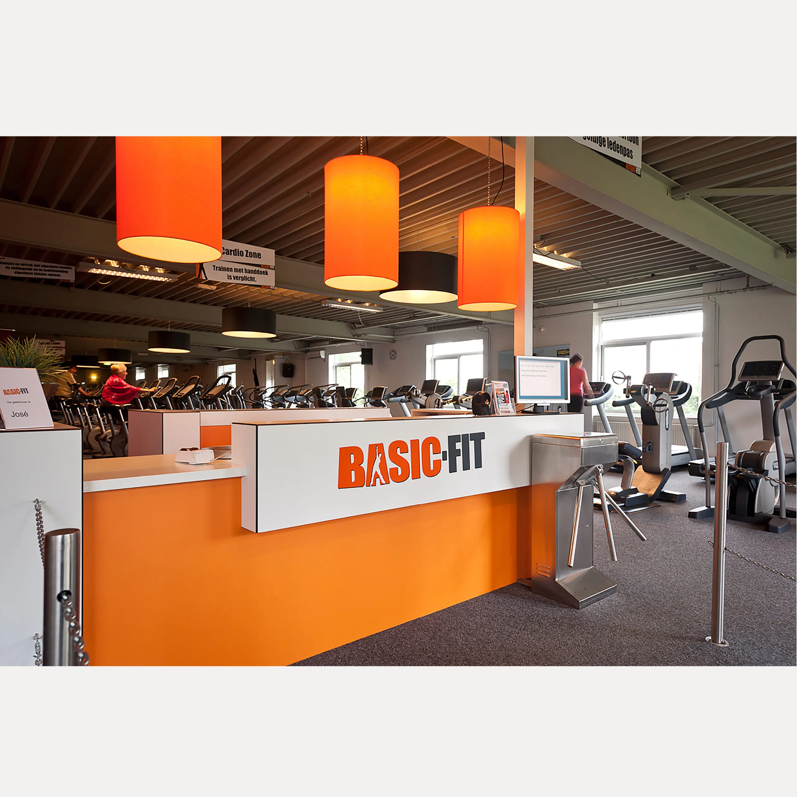 De balie van de BASIC-FIT club in Kerkrade met het woordmerk op het witte paneel en oranje lampen erboven. Clubstyling en logo-ontwerp door Fitbrand.
